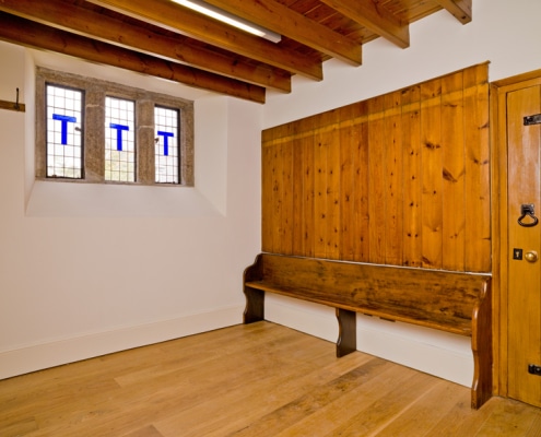 A wooden bench positioned against a wall with a large wooden panel next to a door. Three windows on the adjacent wall with exposed wooden beams on the ceiling.